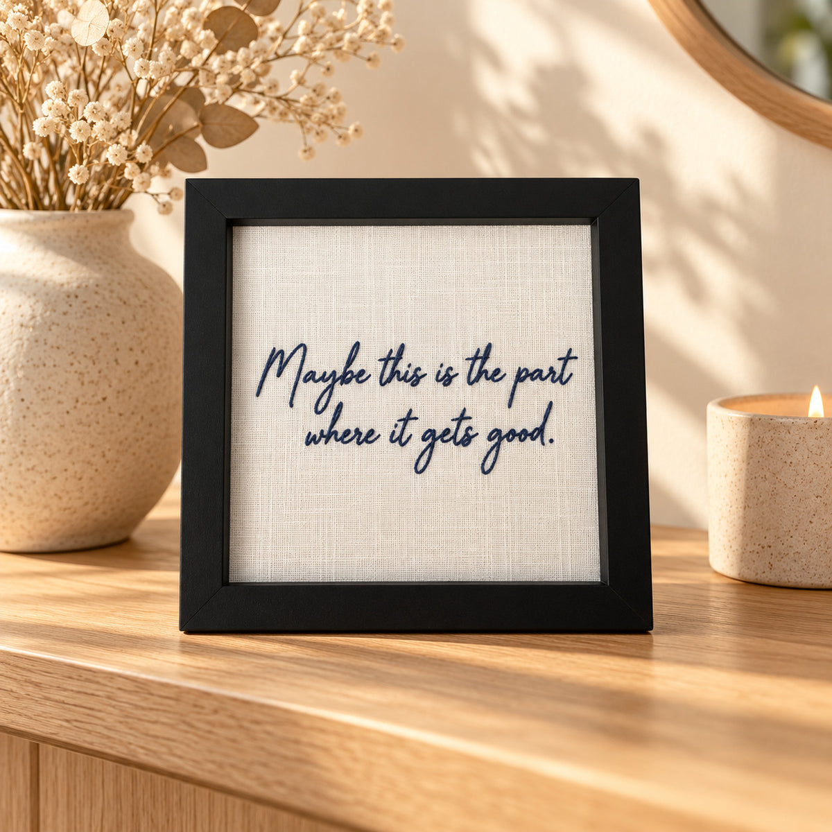 Framed navy embroidered quote 'Maybe this is the part where it gets good' displayed on a wooden sideboard