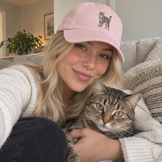 Woman wearing a pastel pink personalised cat portrait embroidered cap with a full-body tabby cat design in a cosy UGC photo